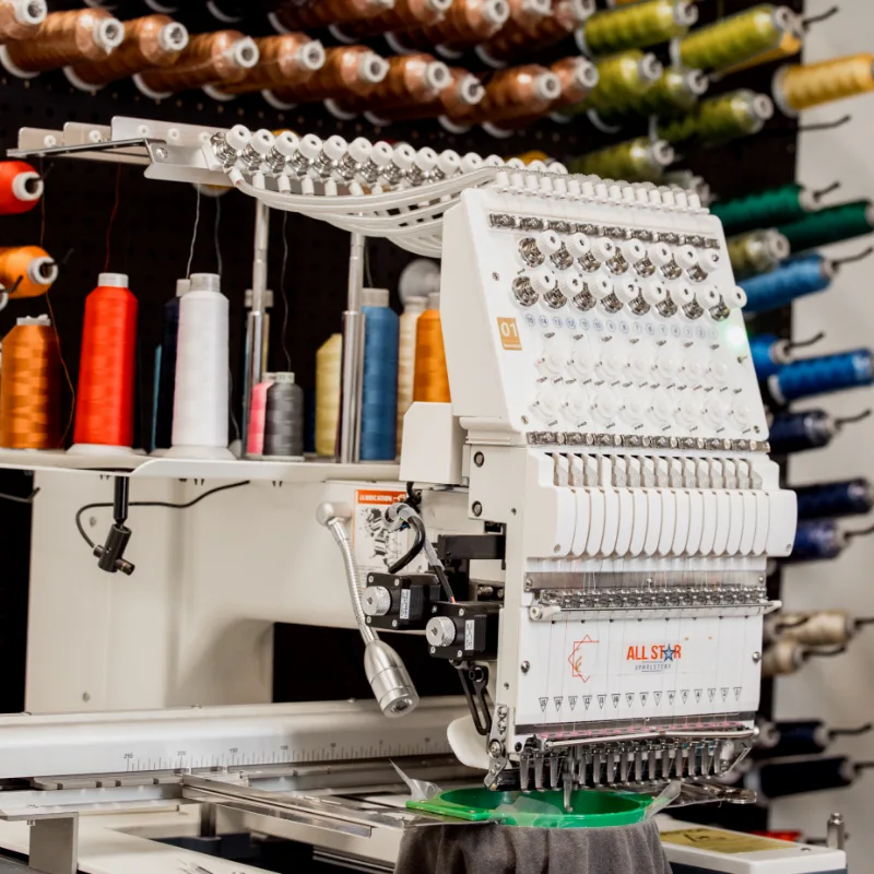 Industrial embroidery machine with All Star Upholstery branding on the side, surrounded by colorful thread spools in the background