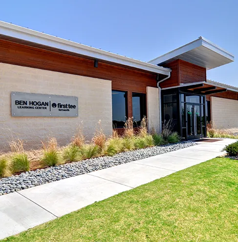 Ben Hogan Learning Center exterior at Squaw Creek Golf Club with a metal sign reading 'Ben Hogan Learning Center | First Tee Fort Worth' mounted on the stained wood and beige stucco facade, with a landscaped pathway and ornamental grasses
