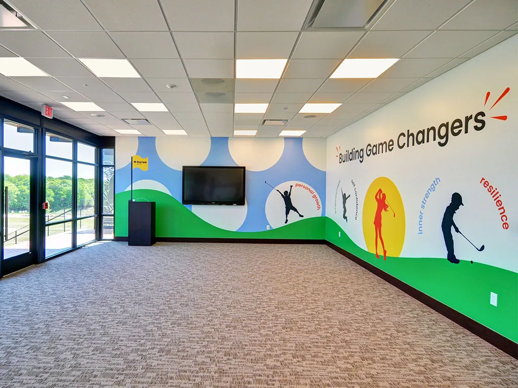 Ben Hogan Learning Center main classroom with a wall-mounted TV, large blue First Tee dot pattern, glass windows overlooking the golf course, and the 'Building Game Changers' silhouette mural