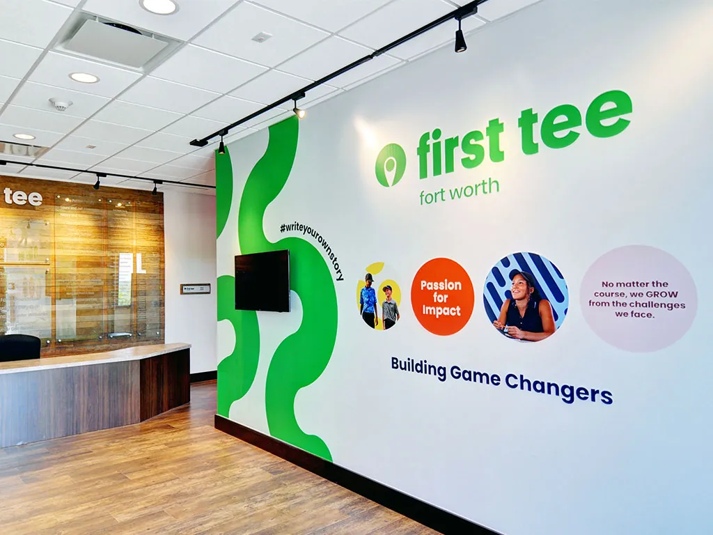 First Tee Fort Worth branded wall at the Ben Hogan Learning Center with the '#writeyourownstory' callout, 'Passion for Impact' circle, member portrait, and the 'Building Game Changers' tagline
