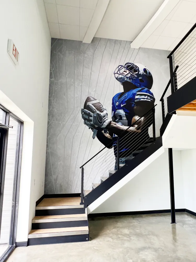 Large-scale vinyl wall graphic of a football player by the office staircase, turning the Champion Sports Construction workspace into a sports facility showroom