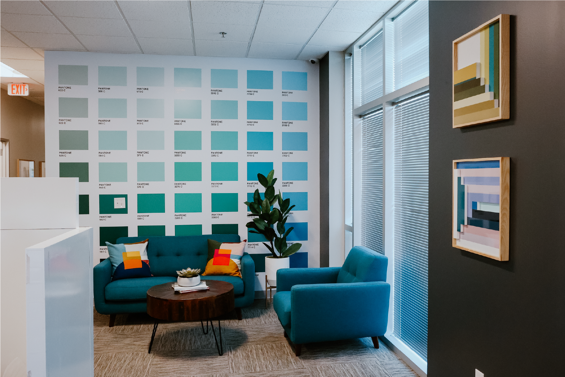 Office sitting area with a wall of Pantone color swatches in teal, aqua, and blue, a teal velvet sofa, accent chair, round wood coffee table, and framed abstract art