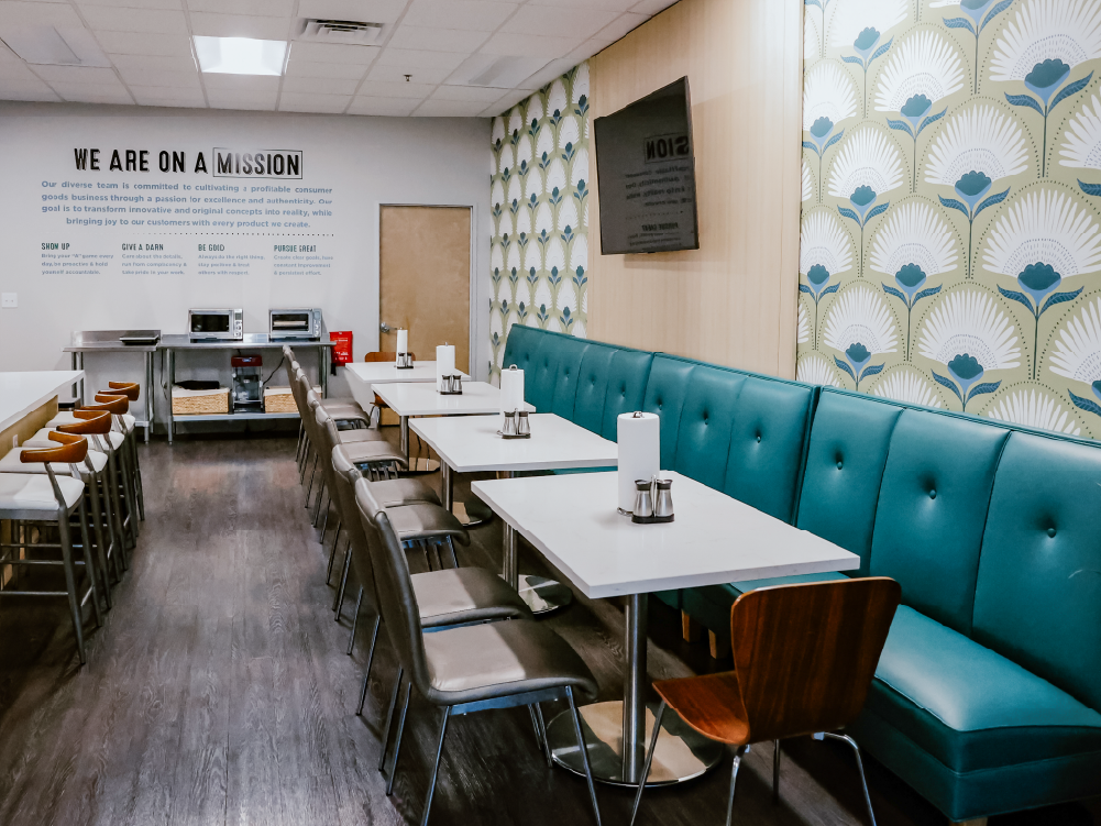Creative Manufacturing breakroom with a 'We Are On A Mission' wall installation, dining tables, and teal upholstered booth seating against magnolia bloom wallpaper
