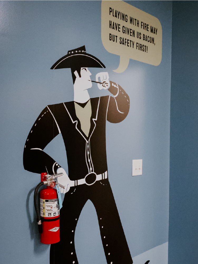 Custom vinyl wall mural of a cowboy holding a fire extinguisher with a speech bubble reading 'Playing with fire may have given us bacon, but safety first!'