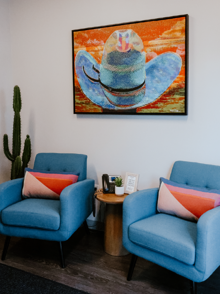 Office seating nook with two teal upholstered armchairs, a tall cactus, and a framed cowboy hat painting on the wall