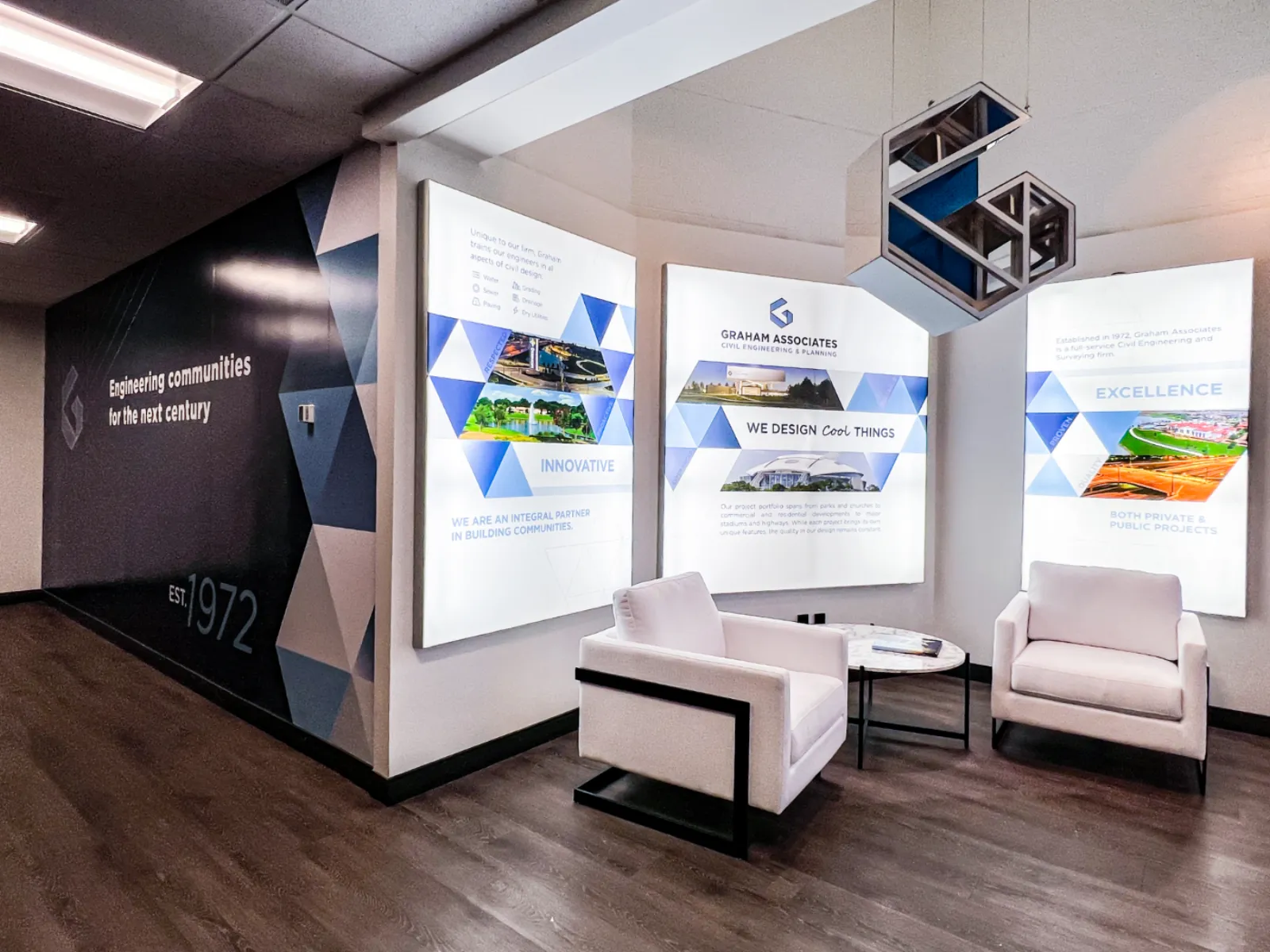 Graham Associates office lounge with three backlit brand display panels, hanging geometric metal sculptures, and a navy accent wall reading Engineering communities for the next century