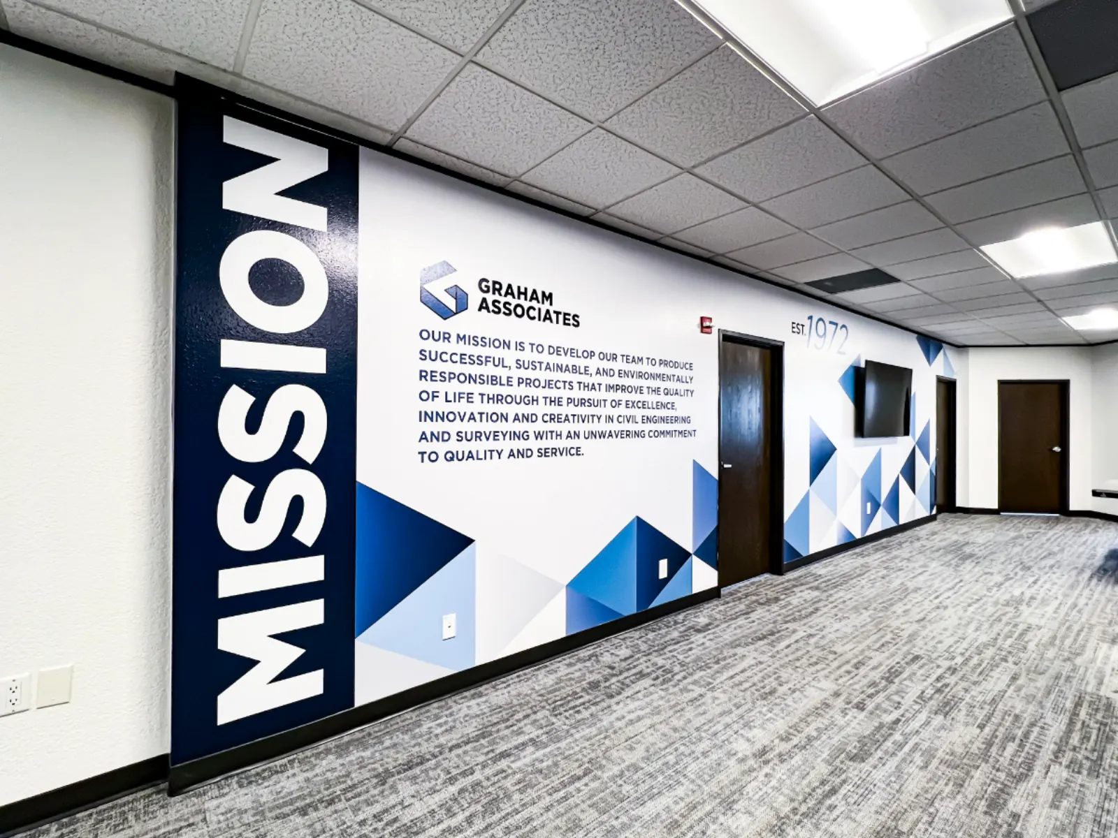 Graham Associates MISSION wall with large vertical typography, the firm's mission statement, the new G mark, and a layered triangle pattern install
