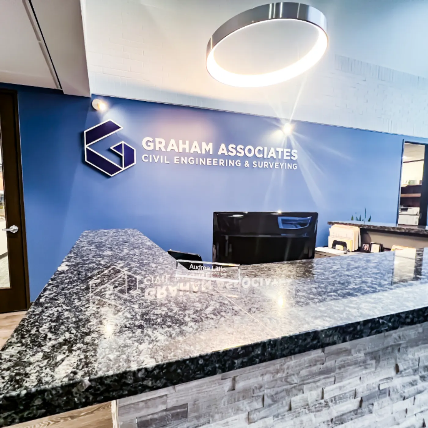 Graham Associates reception desk with a dimensional G mark sign reading Civil Engineering & Surveying on a navy wall above a black-and-gray granite counter