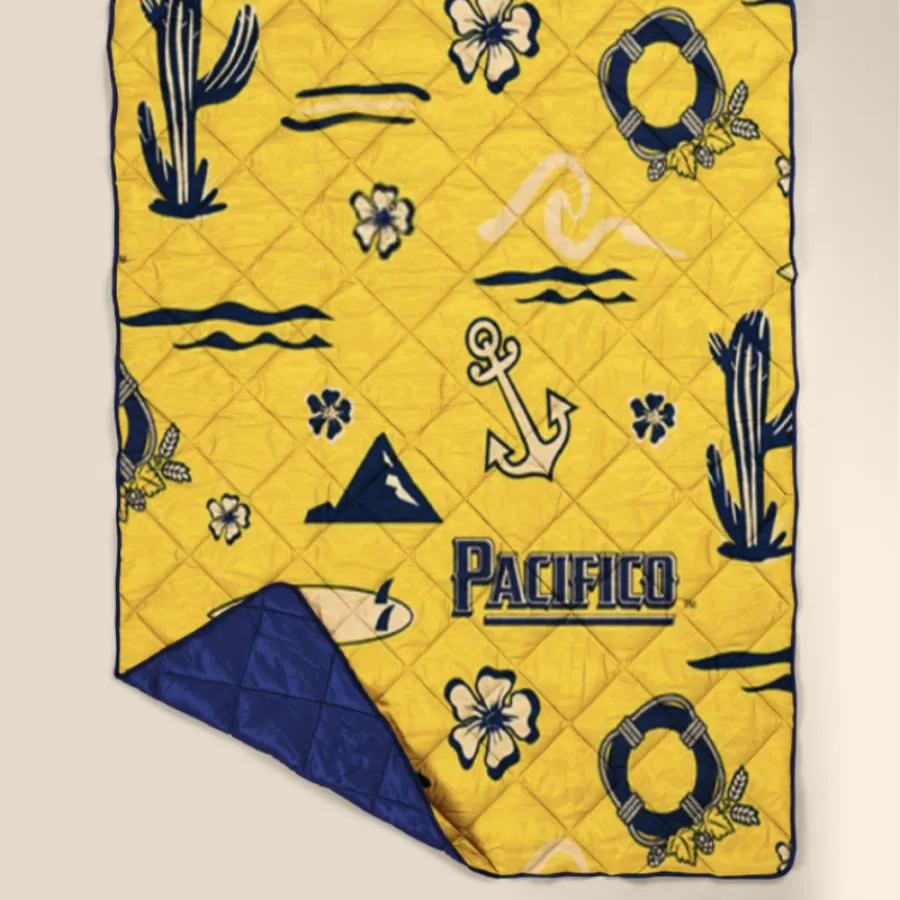 Pacifico-branded patterned quilt blanket in yellow with cacti, anchors, hibiscus flowers, life preservers, and surfboards in navy
