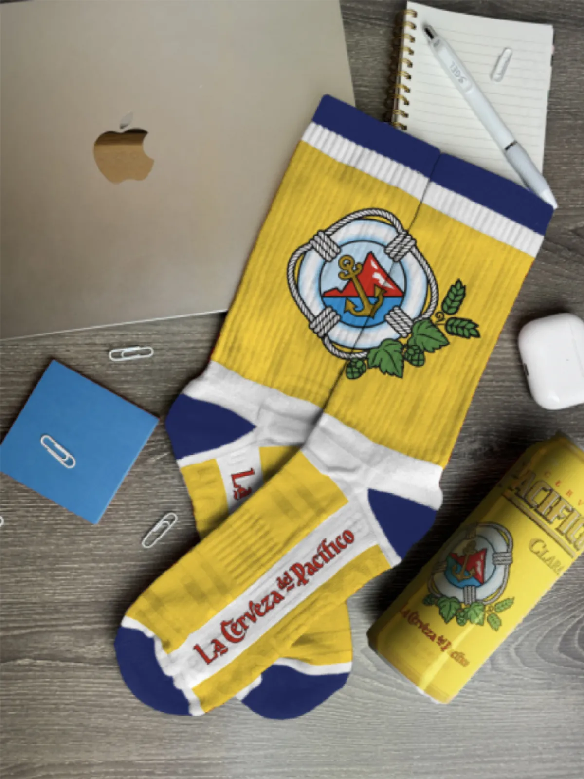 Pacifico-branded socks in yellow and blue with the anchor and Pacifico crest, photographed on a wooden desk with a Pacifico can
