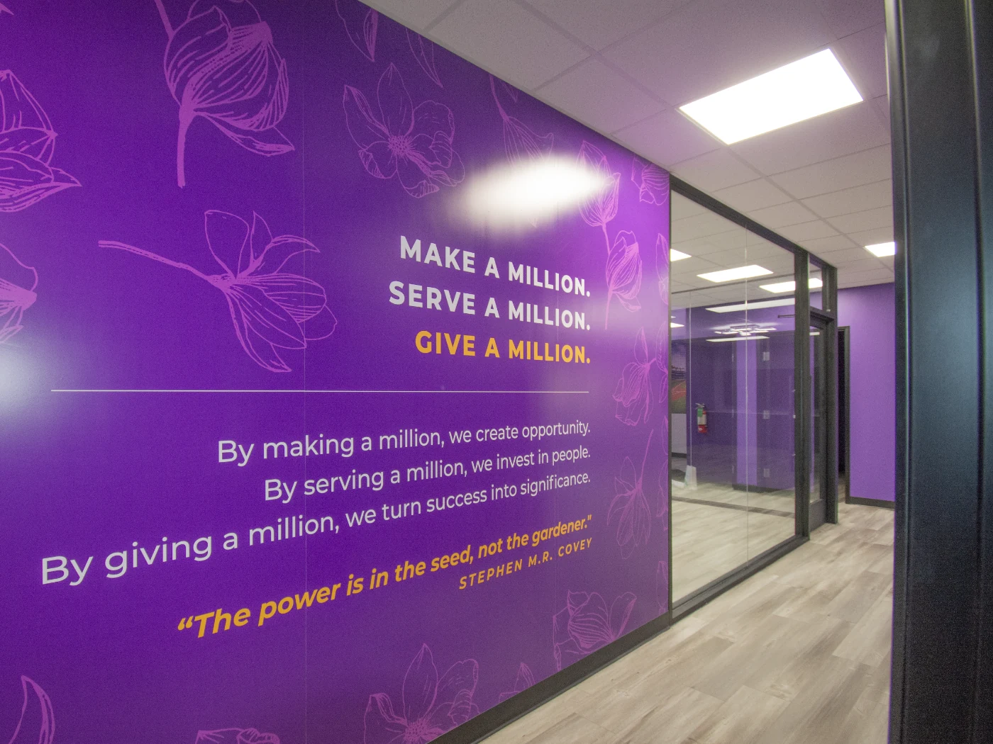 Custom vinyl wall installation reading 'Make a Million. Serve a Million. Give a Million.' over a Stephen M.R. Covey quote, set on a deep purple wall with the magnolia bloom illustration pattern