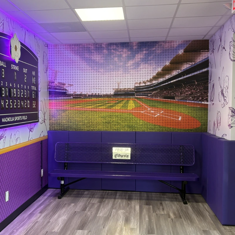 A toddler stands at the Magnolia Fence & Patio office stadium mural, reaching up toward the photographic baseball field wall graphic behind protective netting