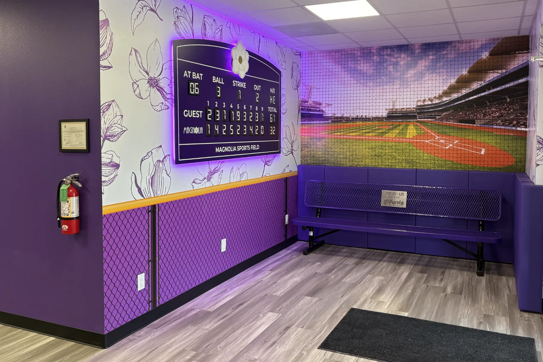 Magnolia Fence & Patio office dugout area with custom backlit scoreboard, stadium photo mural, chain-link fence vinyl wainscot, yellow safety stripe, and purple padded bench seating