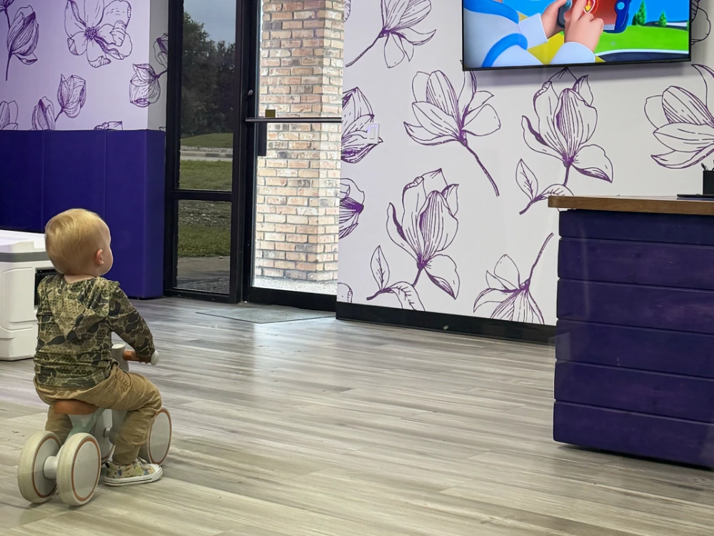 Toddler riding a balance bike past the magnolia bloom wallpaper in the Magnolia Fence & Patio family waiting area with a wall-mounted television