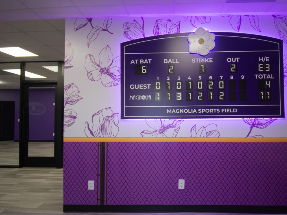 Close-up of the custom dimensional backlit scoreboard with working at-bat counter, magnolia logo, chain-link wainscot, and yellow safety stripe