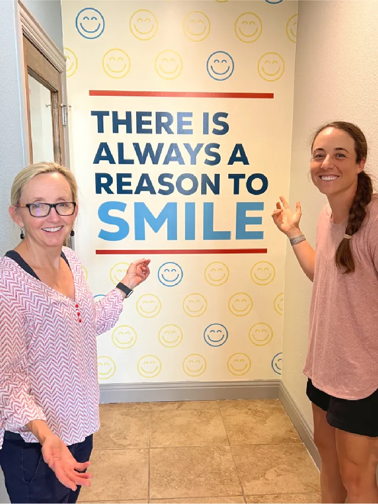 Two Tardy Orthodontics team members standing by the 'There Is Always a Reason to Smile' vinyl wall graphic with a smiley face pattern background
